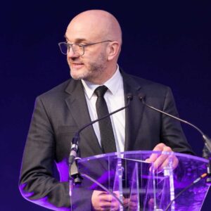 Gareth Healey speaking on stage at the Prolific North Marketing Awards behind a clear lectern in a dark suit and tie.