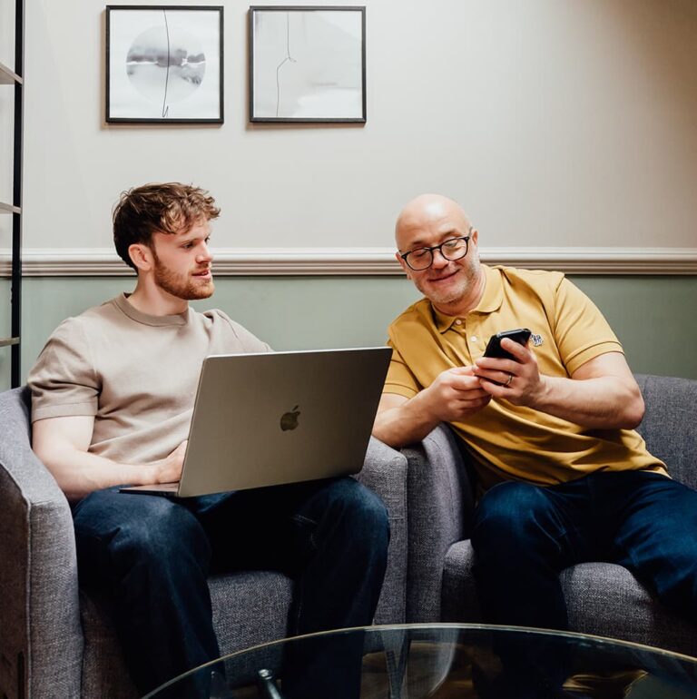 Agents of Change founders Gareth Healey (agency advisor, right) and Callum Healey (AI lead, left) seated in a modern office lounge. This hero image for the About page represents the combination of human strategy and AI intelligence.