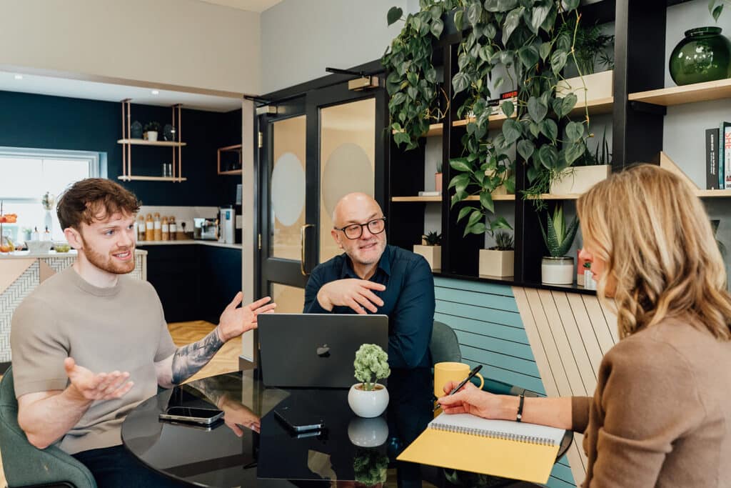 Callum Healey, AI lead at Agents of Change, in a consultation session discussing AI activation outcomes. He is wearing a beige t-shirt and seated at a table, explaining the 90-day roadmap for structured AI implementation to a marketing agency founder.
