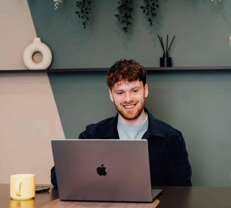 Callum Healey, AI Lead at Agents of Change, smiling while working on a laptop at a desk with a yellow mug. This image is featured on the contact page for founders who prefer to start their enquiry by email.