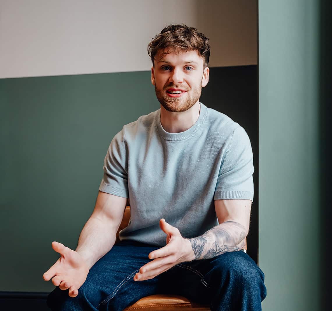 Callum Healey, AI lead at Agents of Change, gesturing while explaining AI activation strategies. He is wearing a light blue t-shirt and seated in a modern office with a green wall. This is the hero image for the AI Activation consultancy service page.