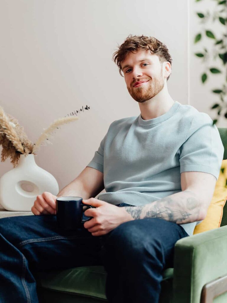 Callum Healey, AI lead at Agents of Change, sitting in a green armchair and smiling at the camera. He is wearing a light blue knitted t-shirt and holding a dark mug. This is the primary bio image for Callum Healey on the About page.