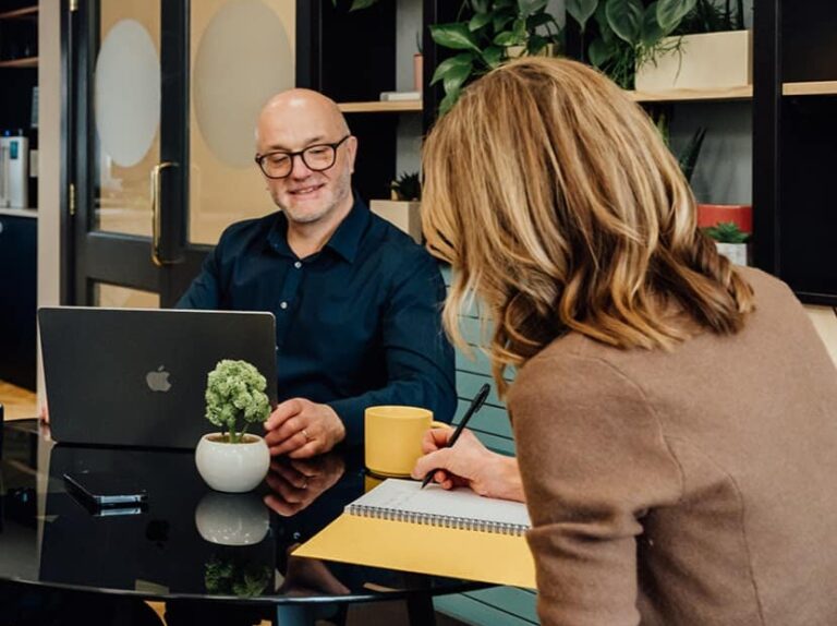 Agency advisor Gareth Healey reviewing data on a laptop during a private consulting session with an agency founder. Gareth is smiling and wearing a navy button-down shirt in a bright, modern office with green plants in the background.