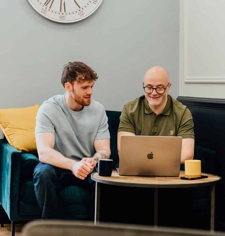 Agents of Change founders Gareth Healey (agency advisor, right) and Callum Healey (AI lead, left) sitting on a navy sofa reviewing a laptop. This image is placed adjacent to the book a discovery call section for marketing agency founders.