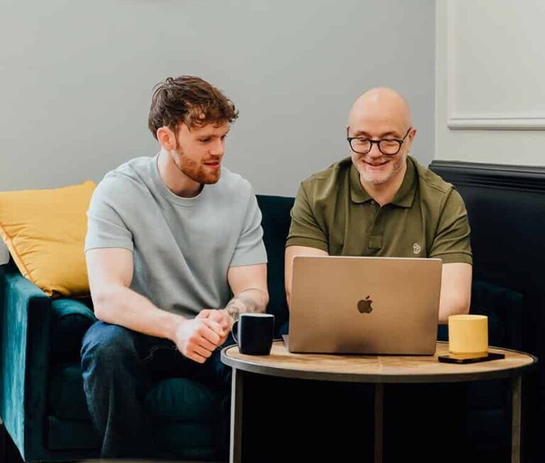 Agents of Change founders Gareth Healey (agency advisor, right) and Callum Healey (AI lead, left) sitting on a navy sofa reviewing a laptop. This image is placed adjacent to the book a discovery call section for marketing agency founders.