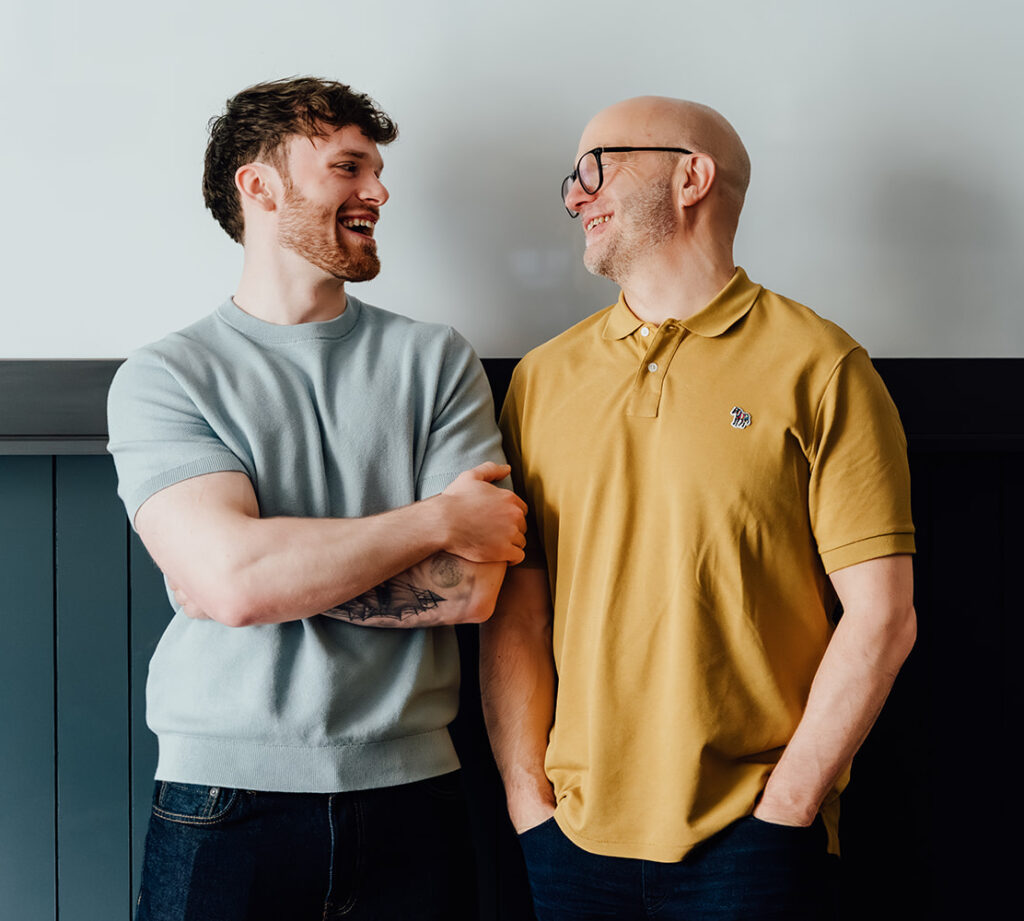 Agents of Change founders Gareth Healey (right, in mustard polo) and Callum Healey (left, in light blue) laughing together, hands in pockets. This image captures the personal partnership and unique consultancy model they offer to marketing agencies.