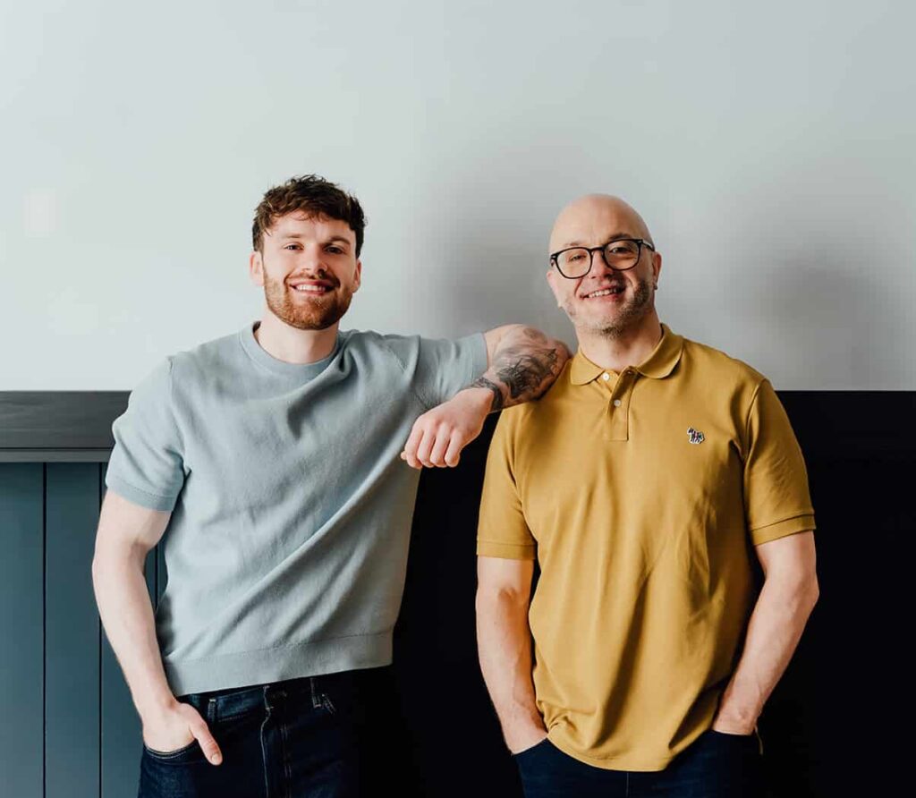 Agents of Change founders Gareth Healey (right, yellow polo) and Callum Healey (left, light blue t-shirt) leaning against a modern dark wall. Gareth Healey is an expert agency advisor, and Callum Healey is the AI Lead helping marketing agencies adopt new technologies.