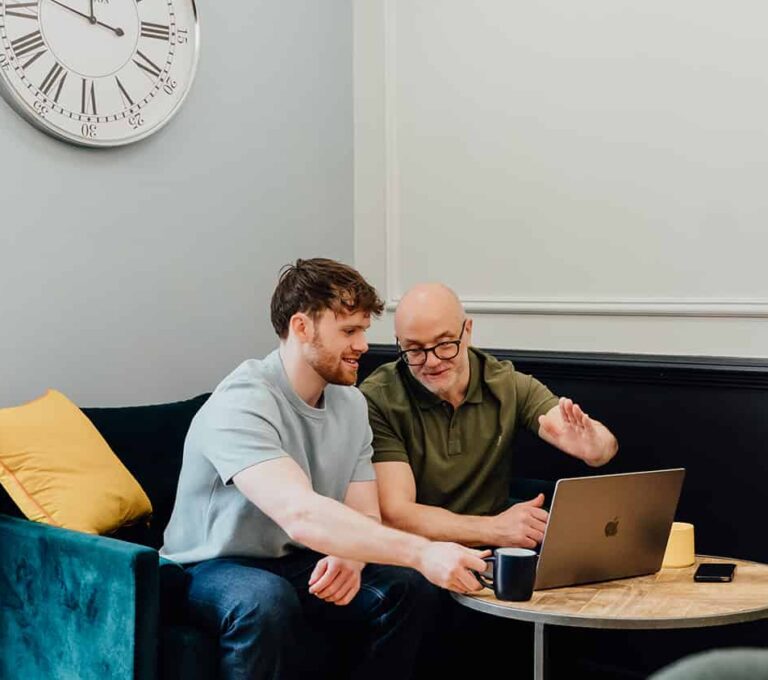 Gareth Healey and Callum Healey of Agents of Change sitting together on a navy sofa, reviewing data on a laptop. Gareth Healey, an agency advisor, is gesturing towards the screen while Callum Healey, the AI lead, looks on.