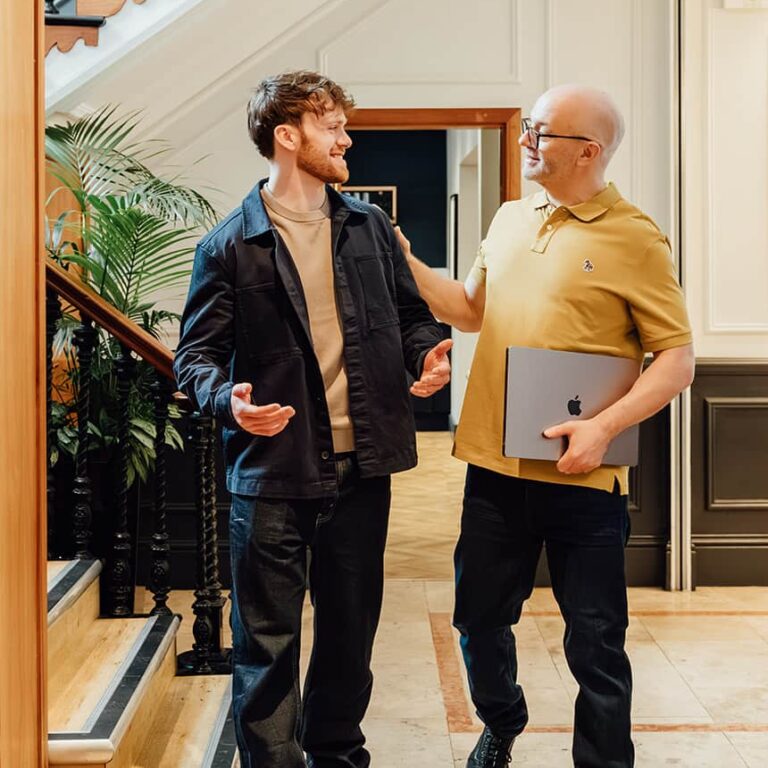 Agency advisor Gareth Healey (right, mustard polo) holding a laptop and talking to AI lead Callum Healey (left, navy jacket) as they walk through a professional office hallway with a staircase. This image represents the collaborative mission of Agents of Change.
