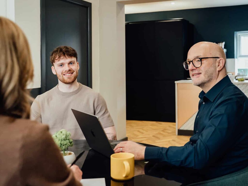 Agency advisor Gareth Healey (right, dark blue shirt) and AI lead Callum Healey (left, beige t-shirt) seated at a table with a laptop, consulting with a client. This image represents the ideal partnership for independent marketing agencies between £400k and £2m revenue.