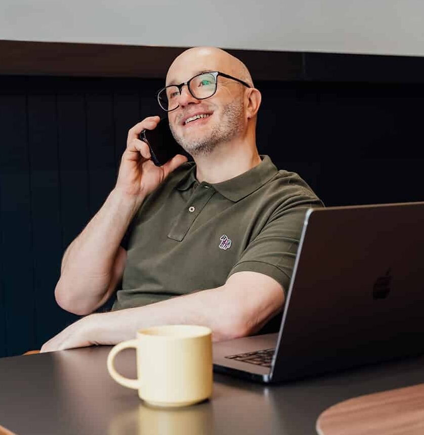 Gareth Healey, an agency advisor with 30 years' experience, smiling while on a business phone call. He is seated at a desk with an open laptop and a yellow mug, wearing a green polo shirt. This image encourages marketing agency founders to get in touch.