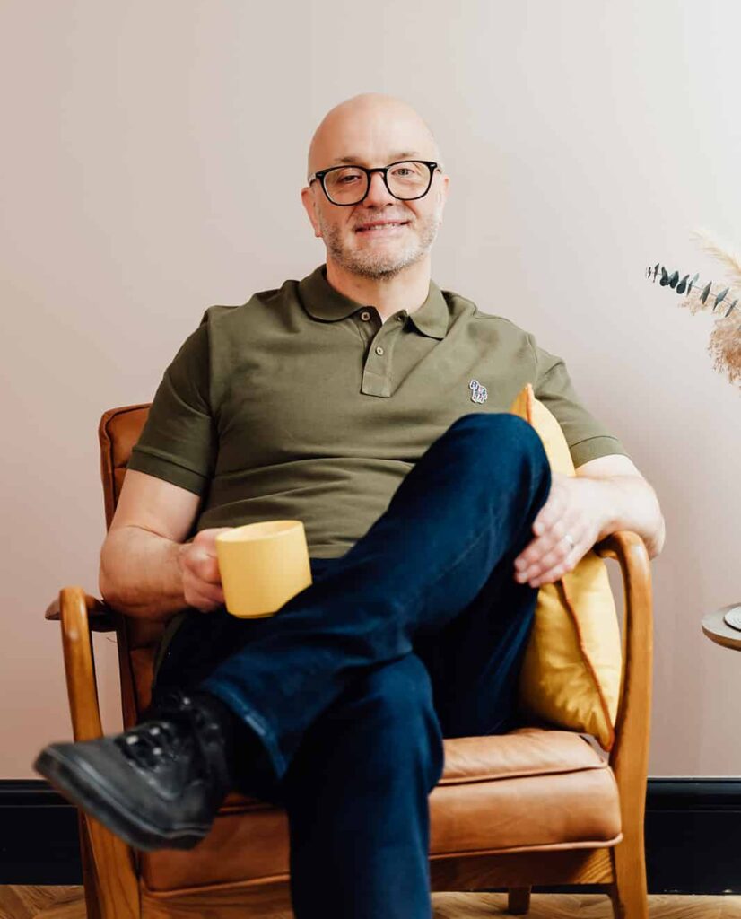 Agency advisor Gareth Healey sitting in a leather armchair, holding a yellow mug and smiling at the camera. He is wearing an olive green polo shirt and glasses. This is the primary bio image for Gareth Healey on the Agents of Change About page.