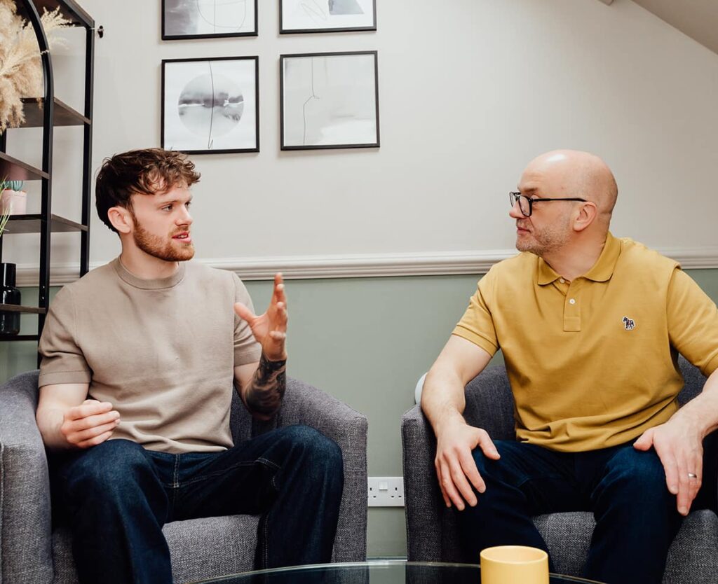 Agency advisor Gareth Healey (right, mustard polo) and AI lead Callum Healey (left, beige t-shirt) seated in grey armchairs, discussing the three core agency objectives: Profit, Growth, and Control. This image illustrates the strategic advisory approach of Agents of Change.