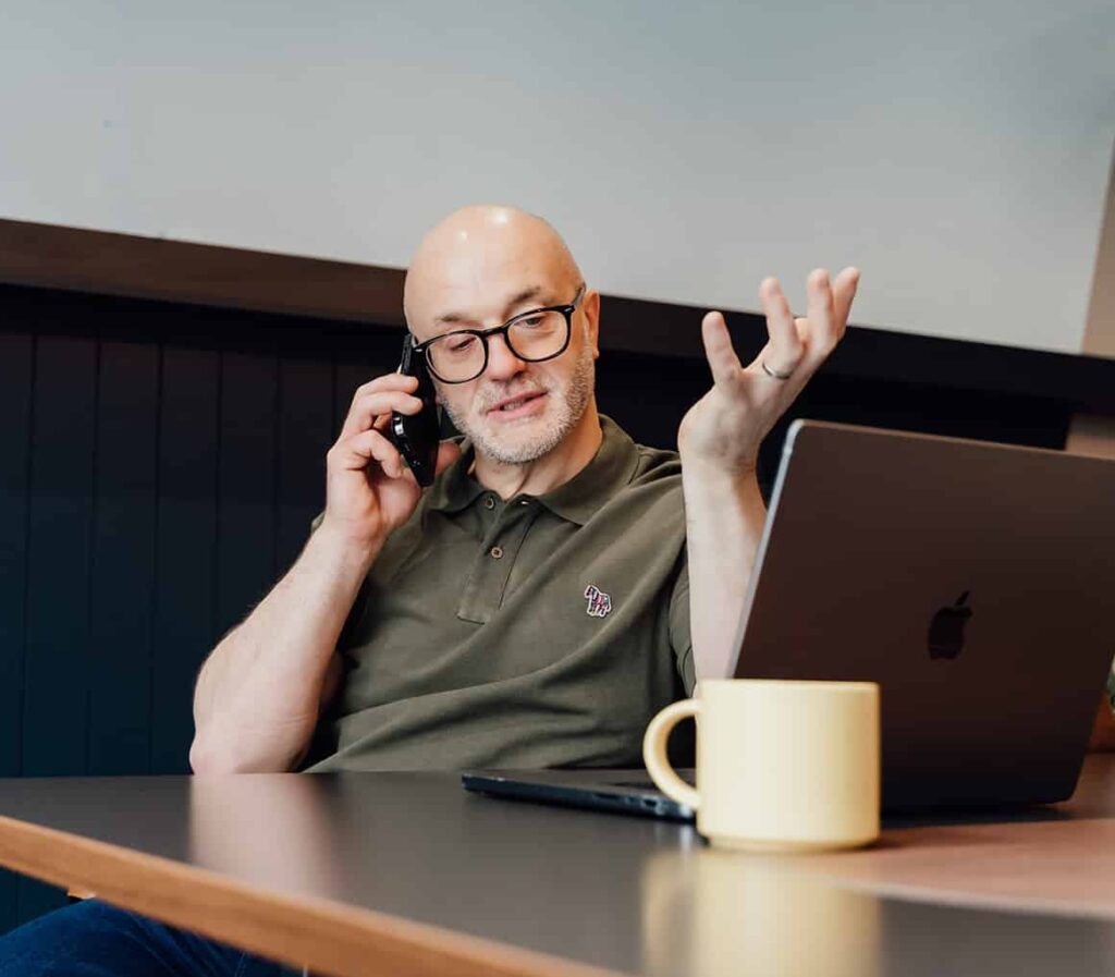 Agency advisor Gareth Healey on a phone call, gesturing with his hand while discussing strategic business constraints. He is seated at a desk with an open laptop and a yellow mug. This image accompanies text regarding agency growth challenges.