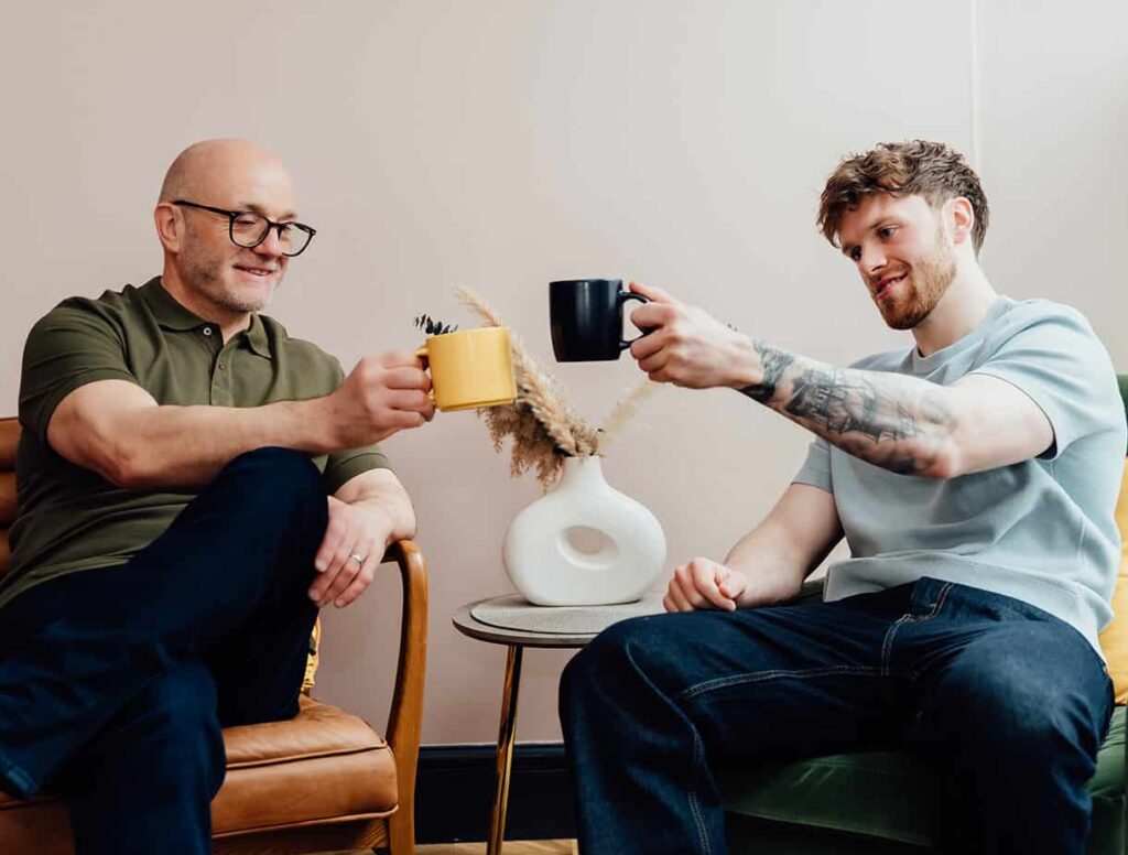 Agents of Change founders Gareth Healey (agency advisor) and Callum Healey (AI lead) clinking their mugs together while seated in leather and green armchairs. This image represents the partnership and founding story of the consultancy.