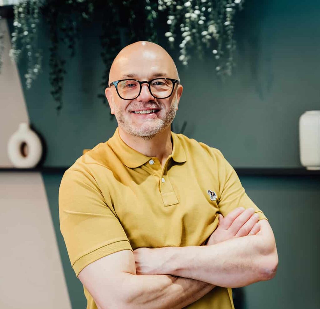 A professional headshot of Gareth Healey, a dedicated agency advisor with 30 years' industry experience. He is smiling warmly, wearing a yellow polo shirt and glasses, standing with arms crossed. This is the hero image for the Agents of Change agency advisory services page.