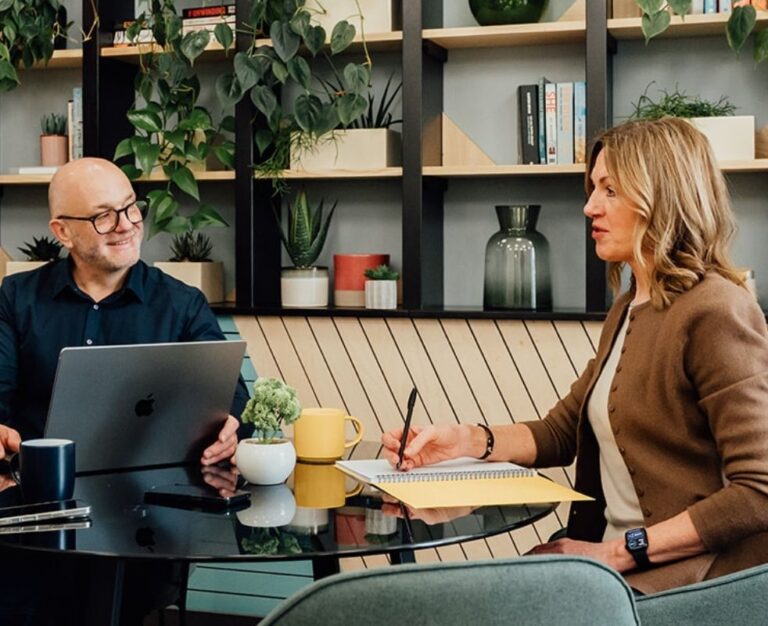 Agency advisor Gareth Healey seated at a round black table with a laptop, listening intently to a female agency founder who is taking notes in a yellow notebook. A yellow mug is visible on the table.