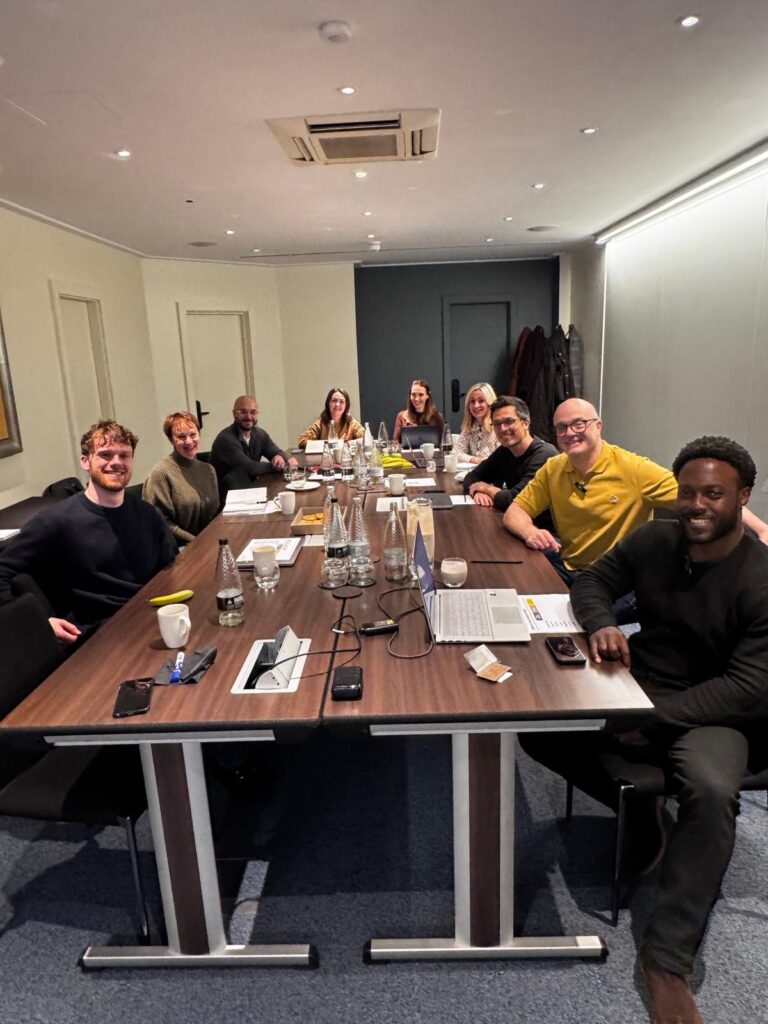 Agency advisor Gareth Healey and AI consultant Callum Healey in a boardroom workshop with a group of marketing agency founders and team members. The group is seated around a long wooden table with laptops and notebooks.