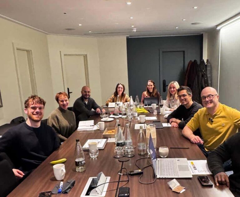 Agency advisor Gareth Healey and AI lead Callum Healey in a boardroom workshop with a group of marketing agency founders and team members. The group is seated around a long wooden table with laptops and notebooks.