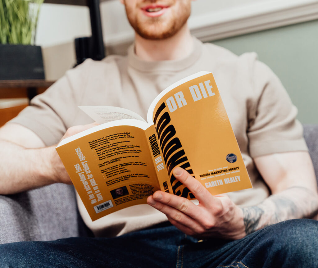 Callum Healey holding a yellow and black book titled 'STANDOUT or DIE' by Gareth Healey. The book discusses making digital marketing agencies stable, desirable, and valuable. This image supports the core beliefs of Agents of Change.