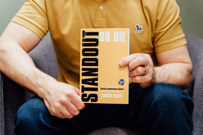 A close-up of agency advisor Gareth Healey holding his book, STANDOUT or DIE. The yellow book cover is prominently displayed, focusing on making digital marketing agencies stable and valuable.